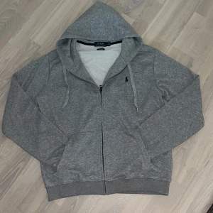 Grå hoodie från Polo Ralph Lauren - Snygg grå hoodie från Polo Ralph Lauren med dragkedja och huva. Klassisk broderad logga på bröstet, två fickor framtill och justerbara snören i huvan. Mjuk bomullsblandning och relaxed fit – perfekt för chill dagar.