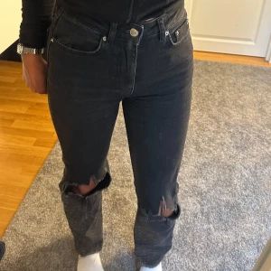Svarta slitna jeans med raka ben - Säljer ett par svarta jeans med raka ben och snygga slitningar vid knäna. Jeansen har klassisk femficksdesign, hög midja och är i ett mjukt denimtyg. Perfekta för dig som gillar en avslappnad och trendig look.