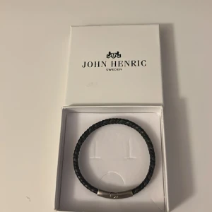 Svart flätat armband John Henric - Stilrent svart flätat armband från John Henric med ett snyggt spänne i silverfärgad metall. Perfekt accessoar för att lyfta din outfit. Kommer i originalask. Oanvänt.