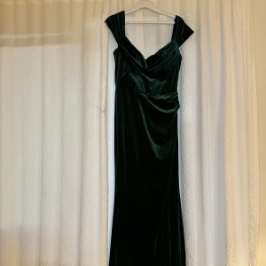 Mörkgrön sammetsklänning med slits - Elegant långklänning i mörkgrön sammet med draperad urringning och breda axelband. Klänningen har en snygg slits framtill och figurnära passform som ger en lyxig känsla. Perfekt för dig som vill sticka ut med en glamorös look.