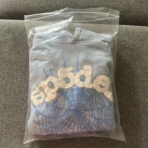 Ljusblå hoodie med tryck - Ljusblå hoodie från  i 100% bomull med coolt vitt och blått spindelnätsmönster och broderad logga framtill. Klassisk huva och mjukt material, perfekt för dig som gillar streetwear och unika prints.