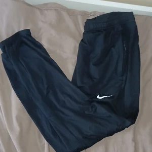 Svarta mjukisbyxor från Nike - Svarta mjukisbyxor från Nike med klassisk vit swoosh-logga på benet. Byxorna har resår i midjan och är tillverkade i ett mjukt material som passar perfekt för chill eller träning. Enkel och stilren design som funkar till det mesta.