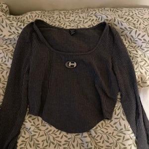 Randig långärmad topp med brosch - Säljer en mörkgrå långärmad topp. Tröjan har en djup, rundad urringning och en snygg brosch med glittrande detaljer framtill. Storleken är xs men känns tight, perfekt för en trendig look.