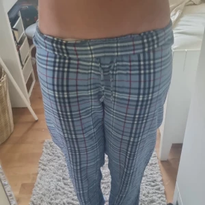 Blårutiga byxor från Gina Tricot - Säljer ett par blåa byxor från Gina Tricot med rutig design i blått, vitt och rött. De har resår och snörning i midjan för skön passform. Perfekta för chill dagar hemma eller som mjukisbyxor. Materialet är mjukt och lätt, vilket gör dem riktigt bekväma.