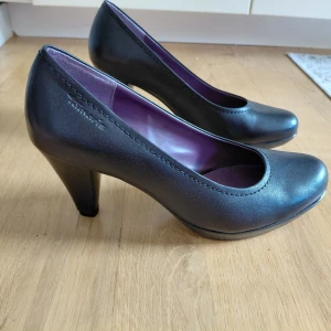 Svarta pumps från Tamaris i SKINN ! - Klassiska svarta pumps från Tamaris i äkta skinn med rund tå och klack på 8cm. Insidan är lila och har snyggt mönster. Perfekta för dig som gillar stilrena och tidlösa skor med en bekväm passform. Sulan utan anmärkning.