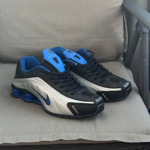 Nike Shox Ultra Blue sneakers  - Nike Shox sneakers med futuristisk vibe i svart, blått och silver. Skorna har den ikoniska fjädrande Shox-sulan, svart ovandel med blå detaljer och silvriga paneler. Snygg snörning och rund tå. Perfekta för dig som vill sticka ut med en sportig och modern look.