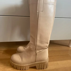 Beiga höga boots med chunky sula - Snygga höga boots i beige med grov, chunky platt sula och rund tå. Skorna är i ett slätt syntetmaterial som ger en clean look. Perfekta för att lyfta din outfit med en trendig touch.