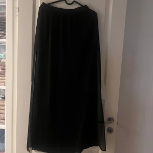 Svart långkjol i transparent tyg - Säljer en svart långkjol med flowig och transparent yttertyg samt en kortare underkjol. Kjolen har en enkel och stilren design med resår i midjan, perfekt för dig som gillar en clean och elegant look.