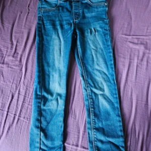 Blå jeans LAB stl 122 - Snygga blå jeans från LAB i storlek 122. Klassisk modell med normal passform och ljusa slitningar framtill. Jeansen har resår i midjan och är tillverkade i hållbar denim. Perfekta för en cool och avslappnad look. Lite slitna framtill, se bild
