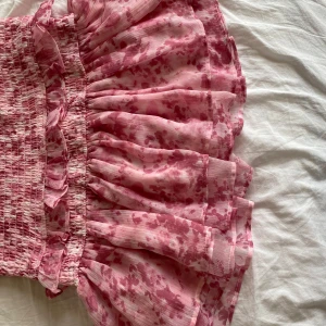 Rosa volangkjol från Gina Tricot - Supersöt kortkjol från Gina Tricot i ljusrosa med mörkare rosa blommigt mönster. Kjolen har smock i midjan, volanger och ett luftigt, lätt tyg som ger en flowy känsla. Perfekt för dig som gillar romantiska vibes och vill sticka ut lite extra.