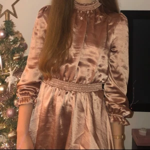 Rosé kortklänning i satin med volang - Superfin kort klänning i skimrande rosa satin med lång ärm, smockad hög krage och midja samt volangdetaljer vid ärmslut och hals. Perfekt för dig som gillar romantisk och trendig stil.