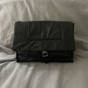 Svart axelväska i skinn med kedja - Snygg svart axelväska i skinn med silverfärgad kedja som axelrem. Väskan har ett lock med spänne framtill och ett stort huvudfack med innerficka som stängs med dragkedja. Perfekt storlek för det viktigaste och stilren design som passar till många outfits. Kan alltid diskutera pris. Skriv för frågor och bilder🥰🥰