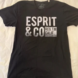 Svart t-shirt från Esprit med tryck - Svart t-shirt från Esprit med stort vitt tryck på bröstet där det står 'ESPRIT & CO REG.TM CALIF.'. Klassisk rund hals och korta ärmar. Perfekt för dig som gillar enkel och stilren streetwear med tydlig logga.
