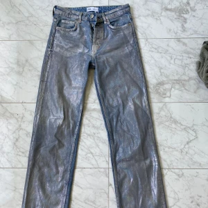 Silvriga straight jeans från Zara - Säljer ett par unika jeans från Zara i storlek 34. De är blå med en metallic silvrig finish som ger en riktigt cool look. Modellen är straight och midwaist med klassiska fem fickor och dragkedja. Perfekta för dig som vill sticka ut lite extra.