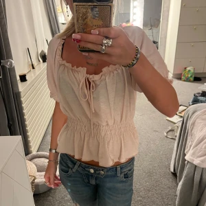 rosa topp  - Supersöt beige topp från Gina Tricot med volangkant, smockad midja och knyt fram. Kan bäras både offshoulder och med puffärm. Tunt, luftigt material som passar perfekt till jeans eller kjol.