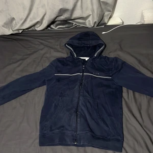 Marinblå hoodie från BOSS med dragkedja - Snygg marinblå hoodie från BOSS med hel dragkedja och huva. Tröjan har en vit linje över bröstet, fickor framtill och BOSS-logga på bröstet. Huva med logotypdetaljer längs kanten. Perfekt för en chill och stilren look.