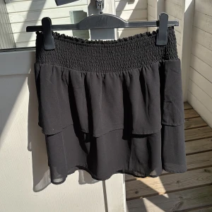 Svart volangkjol från ONLY - Svart kortkjol från ONLY med bred smockad midja och dubbla volanger. Kjolen är gjord i ett lätt och luftigt syntetmaterial som ger en snygg flowig look. Perfekt för dig som gillar en feminin och trendig stil.