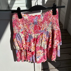 Blommig volangkjol från Kappahl - Supersöt kort kjol från Kappahl med blommigt mönster i rosa, lila, gul och röd. Kjolen har smock upptill och två lager volanger som ger en skön och lekfull vibe. Perfekt för dig som gillar färg och mönster!