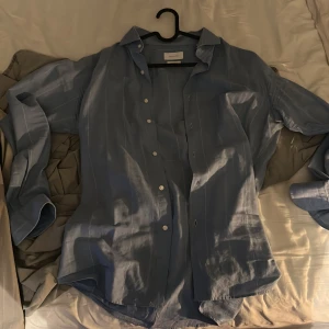 Blå randig skjorta Massimo Dutti - Snygg blå skjorta från Massimo Dutti i bomull och linne. Skjortan har diskreta vita ränder, klassisk krage och knäppning framtill. Perfekt för dig som gillar en clean och stilren look. Långärmad modell med avslappnad passform.