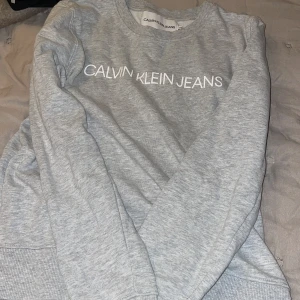 Grå sweatshirt från Calvin Klein Jeans - Snygg grå sweatshirt från Calvin Klein Jeans med vit logotyp framtill. Tröjan har rund hals, ribbade muddar vid ärmslut och nederkant samt långärmad passform. Perfekt för chill dagar och enkel att matcha med jeans eller joggers.