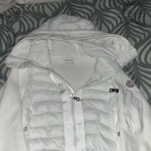 Vit Moncler pufferjacka med huva - Säljer en snygg vit pufferjacka från Moncler med huva och dragkedja framtill. Jackan har ribbade ärmar och sidor i stickad bomull, quiltad front och ikonisk Moncler-logga på ärmen. Perfekt för dig som vill ha en clean och exklusiv look. Det även funkar med QR code på axeln. De ända problemet är att de finns en hål i högra jackan 