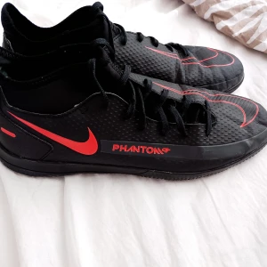 Nike Phantom fotbollsskor svart/röd - Nike Phantom fotbollsskor i svart med röda detaljer och stor Swoosh på sidan och ovansidan. Skorna har snörning, rund tå och en texturerad syntetisk ovandel för extra grepp. Perfekt för dig som vill sticka ut på planen med stil och komfort.Se bilder för skick och storlek. 