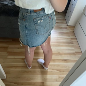 Levi's jeanskjol med slitningar, strl 28 - Säljer en blå jeanskjol från Levi's i klassisk modell med rå kant och slitna detaljer. Kjolen har fem fickor, knapp och dragkedja framtill samt coola fransar längs nederkanten. Perfekt för dig som gillar en avslappnad och trendig stil.