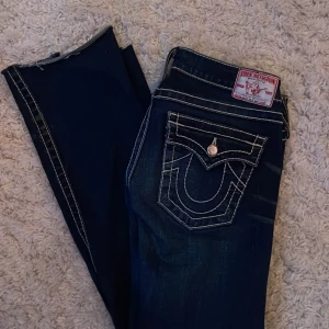 True Religion jeans med vita sömmar - Säljer ett par mörkblå True Religion jeans med snygga vita kontrastsömmar och snygga detaljerade bakfickor med knapp. Jeansen har låg midja och klassisk bootcut passform. Materialet är robust denim och de har coola slitningar framtill. De är ett par 29 i storlek men de sitter som ett bar 26/27 skulle jag säga 