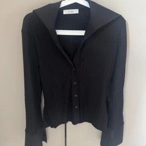 Svart  skjorta från Avora - Snygg svart skjorta från Avora med diskreta ränder och stora kragar. Skjortan har lång ärm, knappar framtill och en lätt croppad passform. Perfekt för dig som gillar stilrena plagg med en twist. Slut online! Helt ny , endast provad 