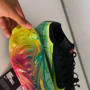 Nike fotbollsskor  vini fly elite - Säljer ett par Nike fotbollsskor med svart ovandel i stickad syntet och snörning. Sidan har ett stort gult Swoosh och textdetaljer i vitt och grönt. Sulan är riktigt fet med regnbågsfärger i gult, rosa, blått och grönt. Perfekt för dig som vill sticka ut på planen.