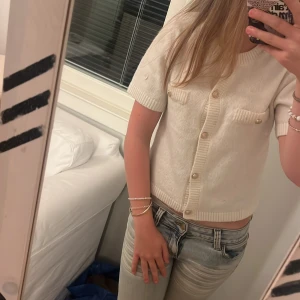 Vit kortärmad stickad blus med knappar - Supersöt vit stickad blus med korta ärmar och guldiga knappar framtill. Två fejkade bröstfickor med dekorativa knappar. Croppad modell med ribbade kanter och mjuk känsla. Perfekt för dig som gillar en clean och chic look.