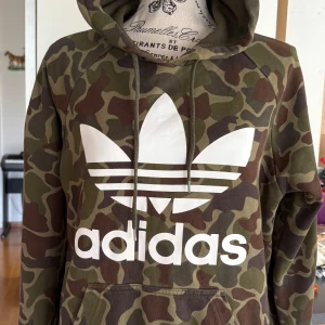 Adidas camo hoodie med logga - Säljer en grön och brun camouflage hoodie från Adidas med stor vit trefoil-logga och text på bröstet. Tröjan har huva med snörning, magficka och långa ärmar. Perfekt för dig som gillar streetwear och sportig stil.