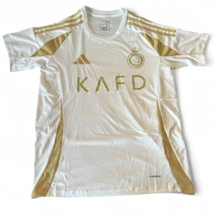 Vit och guld Al Nassr fotbollströja Adidas - Snygg vit fotbollströja från Al Nassr med guldiga detaljer och Adidas-logga. Kortärmad modell med guldiga ränder på axlarna och sidorna, samt klubbmärke och KAFD-tryck på bröstet. Tillverkad i lätt och ventilerande polyester med Aeroready-teknologi.