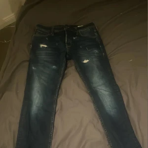 Mörkblå slitna jeans Jack & Jones 32/30 - Säljer ett par mörkblå jeans från Jack & Jones i storlek 32/30. Jeansen har slitna detaljer och en snygg tvätt med ljusare partier på låren. Klassisk femficksmodell med dragkedja och raka ben. Perfekta för en avslappnad streetstil. Egna slitningar