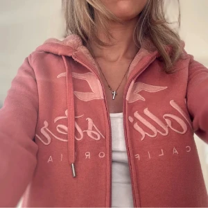 Rosa teddyjacka från Hollister - Mysig rosa teddyjacka från Hollister med huva och dragkedja. Jackan har broderad logga framtill, mjukt teddyfoder på insidan och ribbade muddar. Perfekt för kyliga dagar när du vill vara både bekväm och snygg.