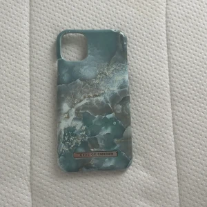  Mobilskal Ideal of Sweden  - Snyggt mobilskal från Ideal of Sweden passar till iPhone 11 med blågrönt marmormönster och detaljer i vitt och guld. Skalets yta har en glansig finish och en metallbricka med loggan längst ner. Passar perfekt för dig som vill ha en stilren vibe på mobilen.