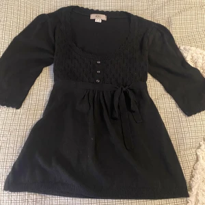 Svart stickad kofta med knyt från bpc - Svart stickad från bpc med snyggt hålmönster upptill, trekvartsärmar och knytband i midjan. Rundad halsringning med dekorativa knappar framtill.