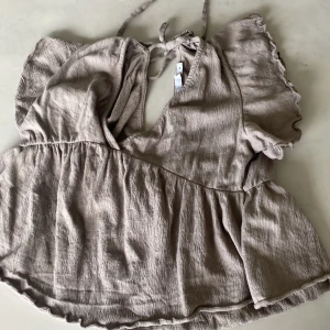 Beige blus med volangärm från 157 - Supersöt beige blus från 157 i storlek XS. Den har korta volangärmar, v-ringning och knytning i nacken. Blusen är luftig med lätt rynk vid midjan och har en flowy, avslappnad passform. Perfekt för dig som gillar en romantisk och chill stil.