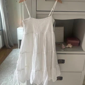 Vit sommarklänning från Gina Tricot - Supersöt vit klänning från Gina Tricot med smock upptill och tunna axelband. Klänningen har en luftig, volangig kjol och är perfekt för varma dagar. Materialet känns lätt och skönt mot huden, och den har en flowy passform som ger en chill vibe.