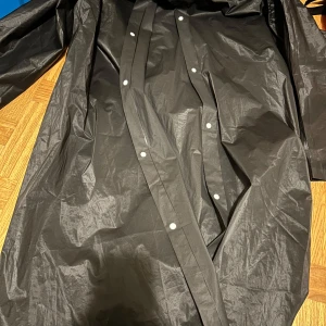 Svart regnjacka med huva - Svart regnjacka med huva och tryckknappar framtill. Jackan är lång och har en clean, minimalistisk look. Perfekt för regniga dagar när du vill hålla dig torr och ändå se stilren ut. Materialet är syntetiskt och vattenavvisande.