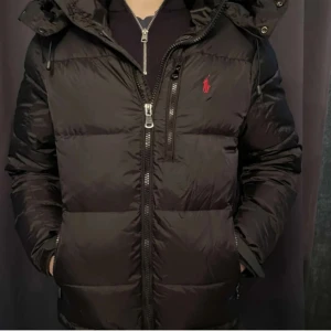 Svart dunjacka från Ralph Lauren - Säljer en svart dunjacka från Ralph Lauren med röd logga på bröstet. Jackan har huva, dragkedja framtill och flera fickor med dragkedja. Puffig modell som håller dig varm och snygg under kalla dagar. Perfekt för dig som gillar stilren och sportig look. Små slitningar finns 
