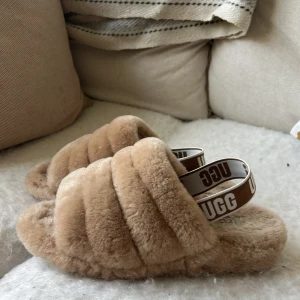 Beige fluffiga sandaler från UGG - Kostade egentligen 800kr.Supermysiga sandaler från UGG i beige fluffigt material med breda band och elastisk rem med UGG-logga. Perfekta för chill hemma eller när du vill ha maxad komfort. Öppen tå och platt sula för skön känsla. Snyggt och trendigt val för dig som gillar cozy vibes.Size är runt 38/39 
