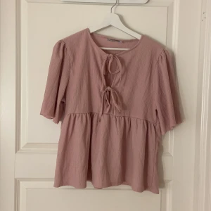 Ljusrosa blus med knyt och volang - Supersöt ljusrosa blus med två knyt framtill och volang nedtill. Blusen har en lätt rynkad material och halvlånga ärmar. Från lager 157 i storlek M men har klippt lite själv i armarna då ja inte tyckte om hur dom såg ut innan 