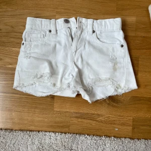 Vita jeansshorts från Zara - Snygga vita jeansshorts från Zara med hög midja och råa, fransiga kanter. Shortsen har slitna detaljer framtill, klassiska fickor och knapp- samt dragkedja. Perfekta för en chill sommarlook och passar dig som gillar streetstyle.