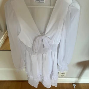 Vit kortklänning från Design By Amor - Supersöt vit kortklänning från Design By Amor i storlek S med långärmade puffärmar, v-ringad hals och knyt framtill. Klänningen har smock i midjan och volangdetaljer längst ner. Kommer tyvärr inte till användning.