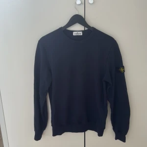 Mörkblå sweatshirt från Stone Island - Snygg mörkblå sweatshirt från Stone Island Junior med klassisk rund hals och långärmad passform. Tröjan har den ikoniska Stone Island-patchen på vänster ärm och är gjord i mjuk bomull. Perfekt för dig som gillar streetwear och vill ha en clean look.