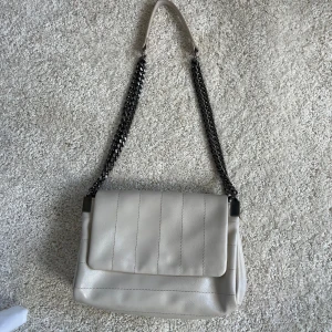 Beige axelväska med kedjerem - Snygg beige axelväska i skinnimitation med coola svarta kedjeremmar. Väskan har lock med diskreta vertikala sömmar och en stilren, rektangulär form. Perfekt storlek för det viktigaste och ger en trendig vibe till din outfit.