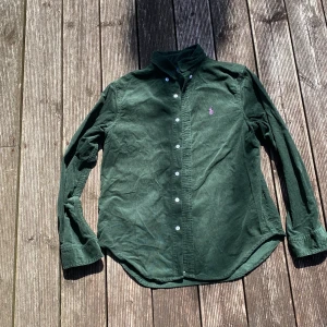 Grön manchesterskjorta Polo Ralph Lauren - Mörkgrön manchesterskjorta från Polo Ralph Lauren med klassisk krage, vita knappar och lila broderad logga på bröstet. Skjortan är långärmad och har en avslappnad passform, perfekt för dig som gillar en stilren men chill look. Det står att den är XL men passformen är som en M.
