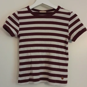 Randig kortärmad topp från Kappahl - Snygg kortärmad topp från Kappahl med breda vinröda och vita ränder. Ribbad och tillverkad i mjuk bomull med lite stretch för skön passform. Storlek 146/152 💕