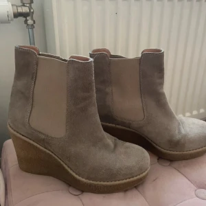 Beige mocka chelsea boots med kilklack - Snygga beige chelsea boots i mocka med bred resår på sidorna och kilklack. Skorna har rund tå och platt sula framtill, perfekt för dig som vill ha både höjd och komfort.